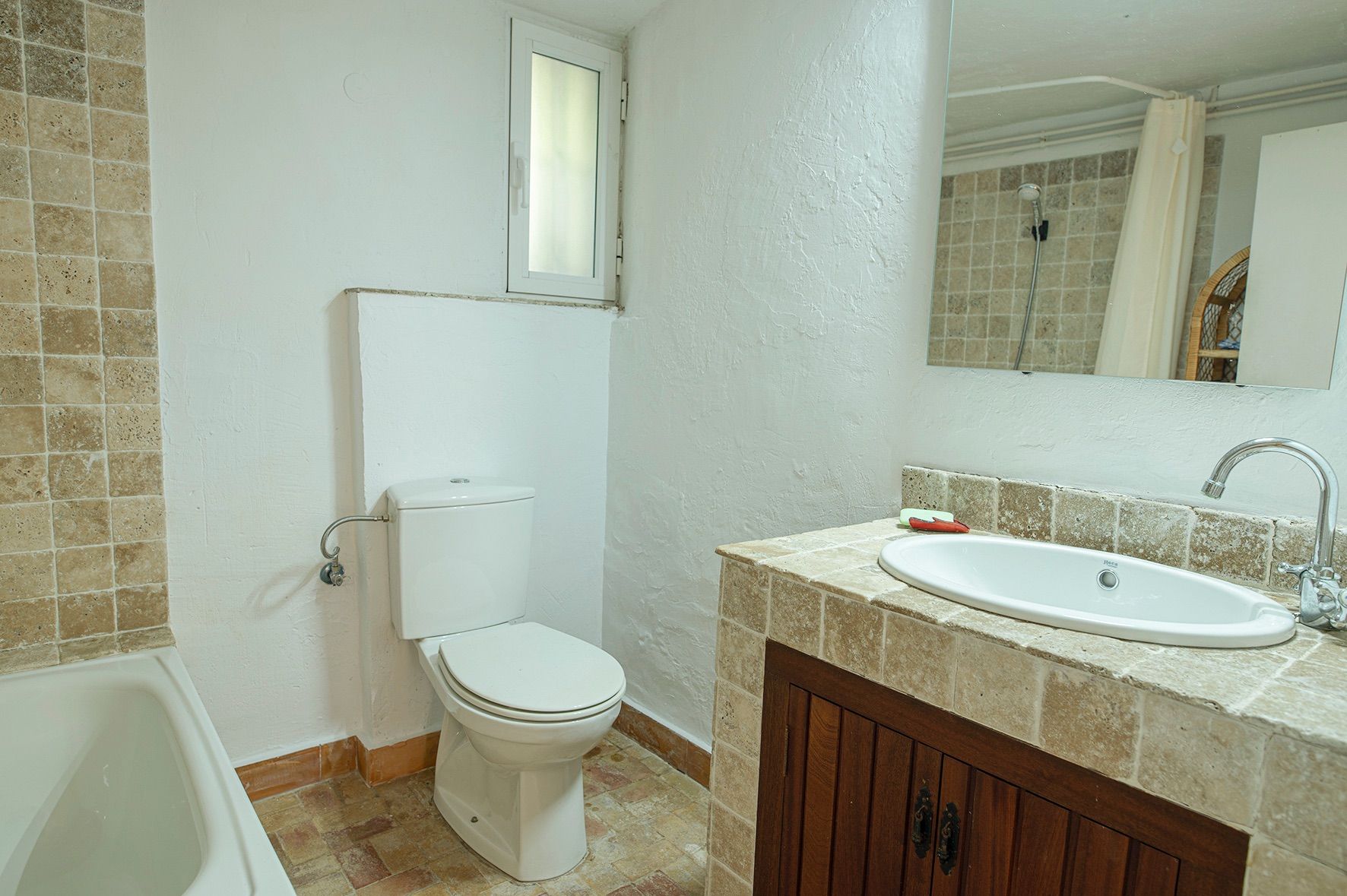 Rustic bathroom featuring a natural stone-tiled vanity with dark wood doors, white ceramic fixtures, and an integrated bathtub.