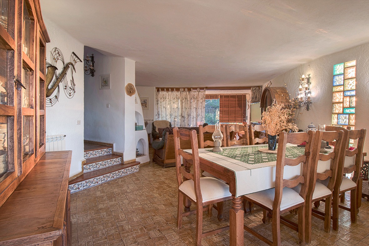 Rustic dining area featuring a wooden table set, terracotta flooring, and stairs with decorative Mediterranean tiles. A colorful glass block window adds natural light.