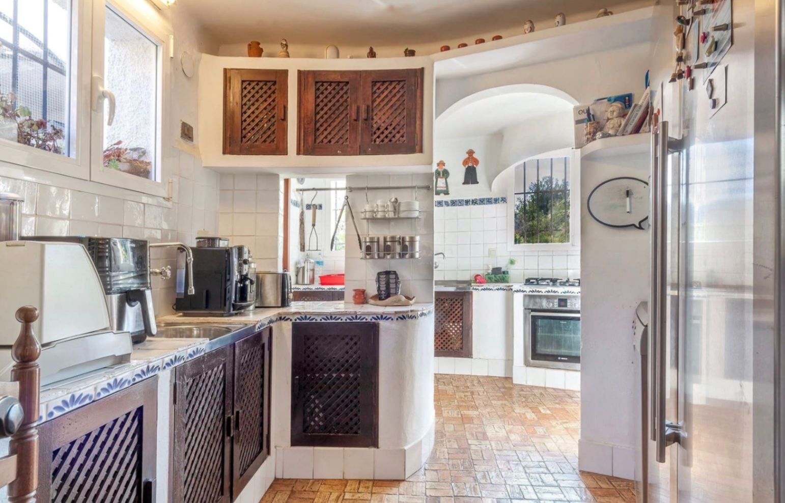 Rustic kitchen featuring dark wood lattice cabinetry, white tiled countertops with blue accents, and traditional terracotta flooring under natural light.
