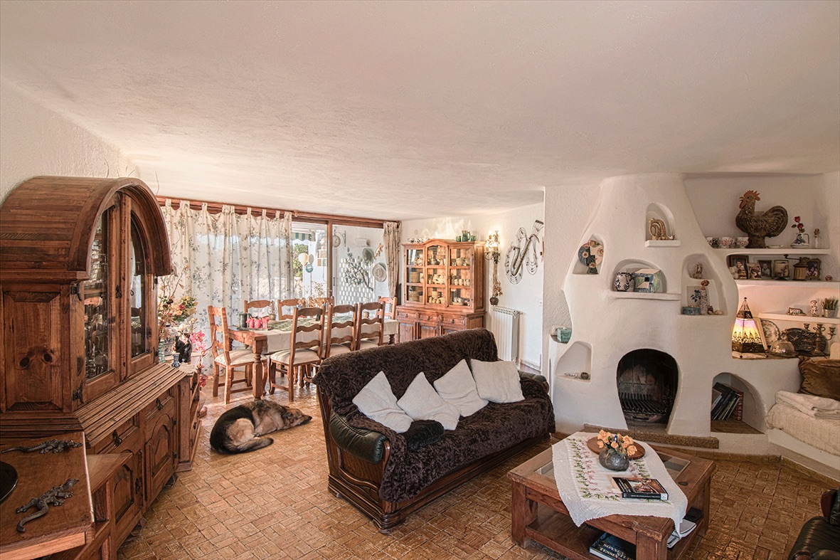 Rustic living and dining area featuring a unique sculpted white fireplace with integrated shelving, solid wood furniture, and traditional terracotta tile flooring.