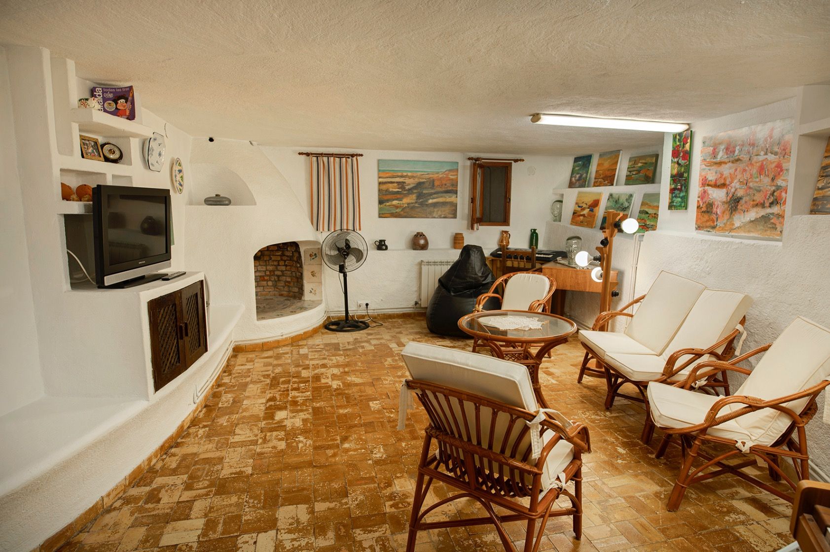 Rustic living area featuring terracotta floors, a built-in fireplace, and whitewashed walls. The space includes bamboo seating and an integrated art studio corner.