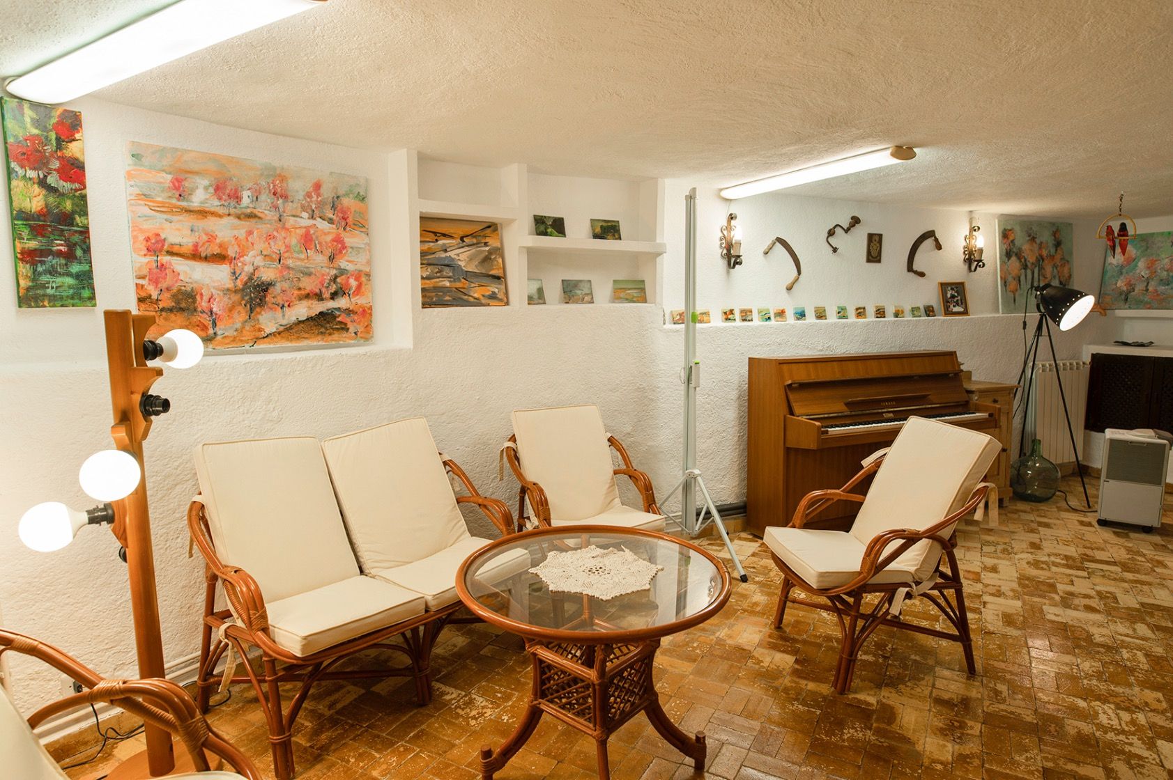 Rustic lounge area featuring rattan seating, an upright wooden piano, and terracotta tile flooring. The textured white walls include built-in shelving and art displays.