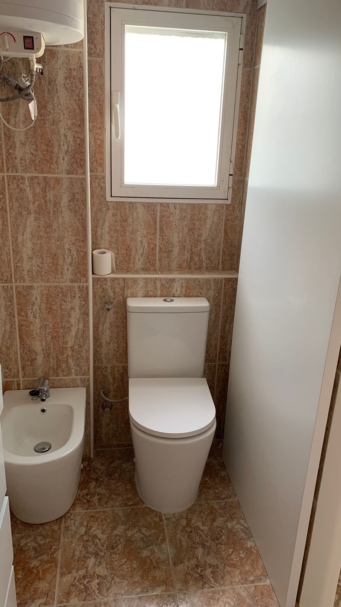 Bathroom with porcelain toilet and bidet, earth-toned tiling, a translucent glass shower screen, and a window for natural light and ventilation.