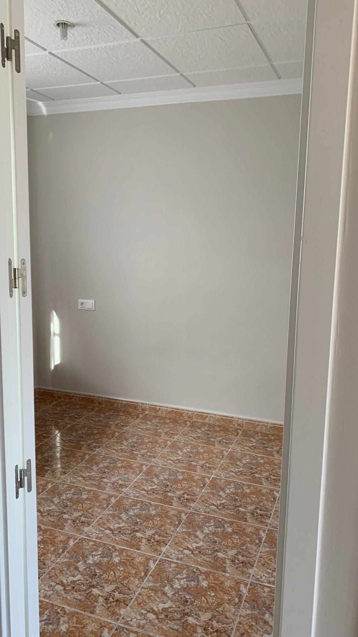 Empty room featuring brown ceramic tile flooring, light grey walls, and a white textured ceiling. Natural light enters from the side, highlighting the clean finishes.