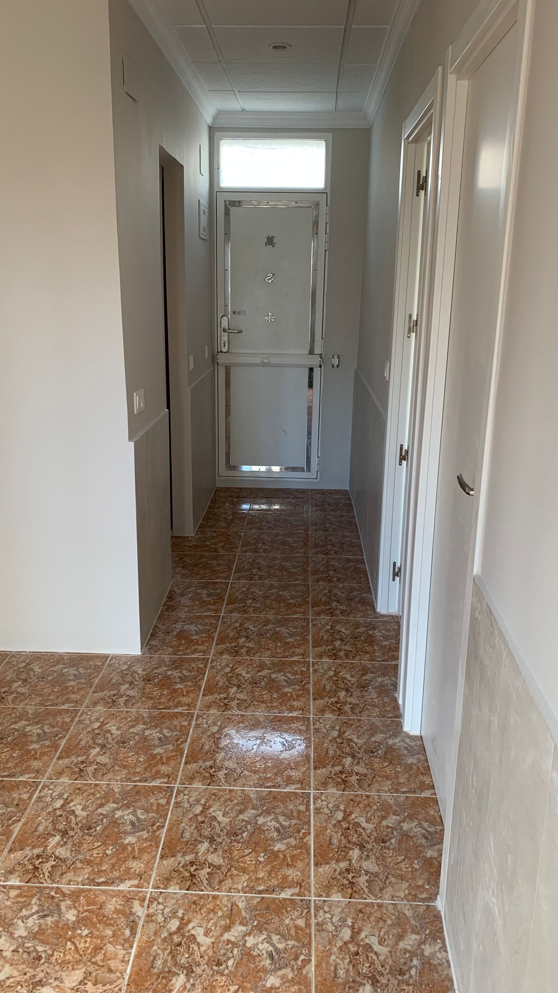 Entrance hallway featuring marbled brown ceramic floor tiles and white walls. A reinforced metal door with a transom window is positioned at the end of the corridor.