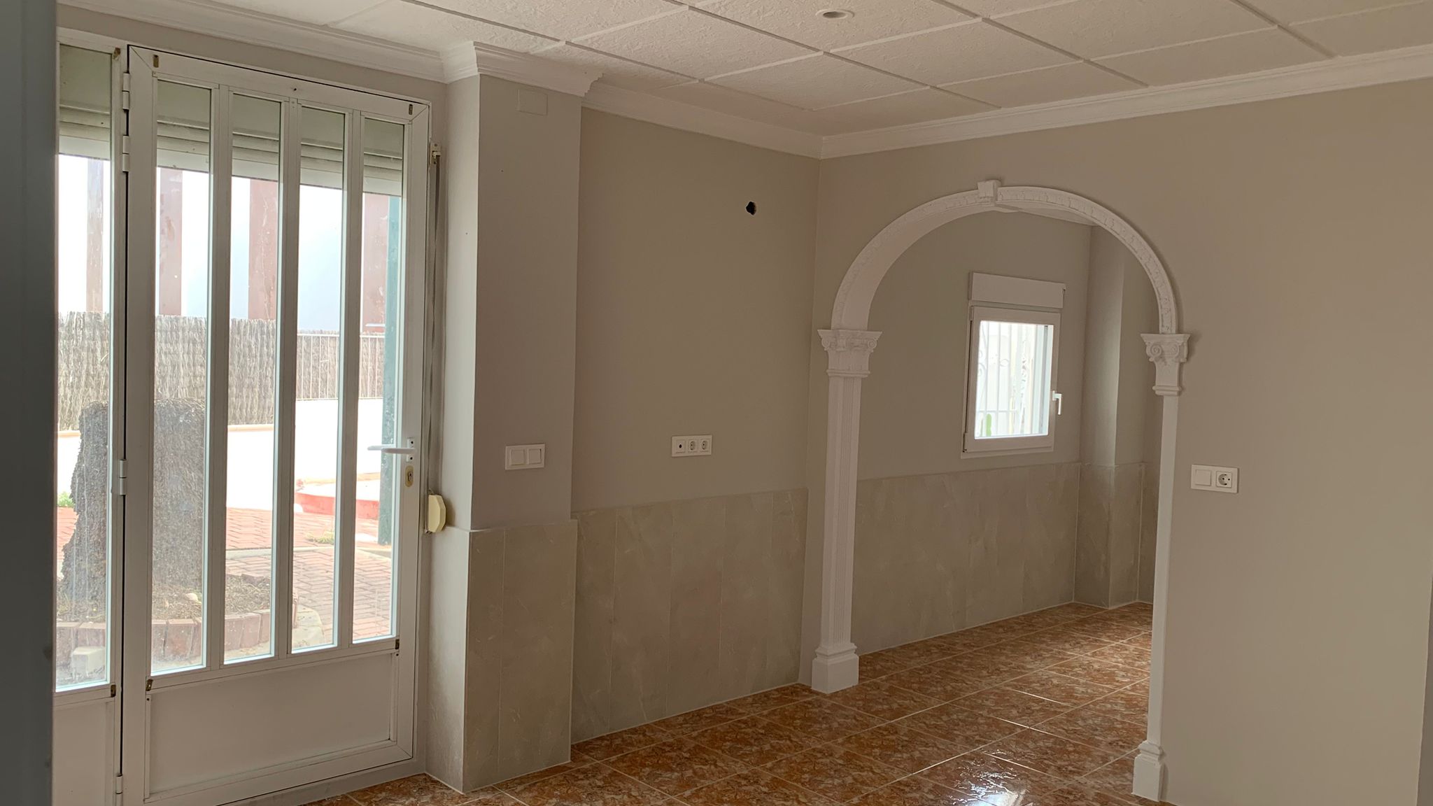 Living area featuring ceramic tile flooring, neutral walls with tiled wainscoting, and a decorative white archway. A glass door provides direct outdoor access.