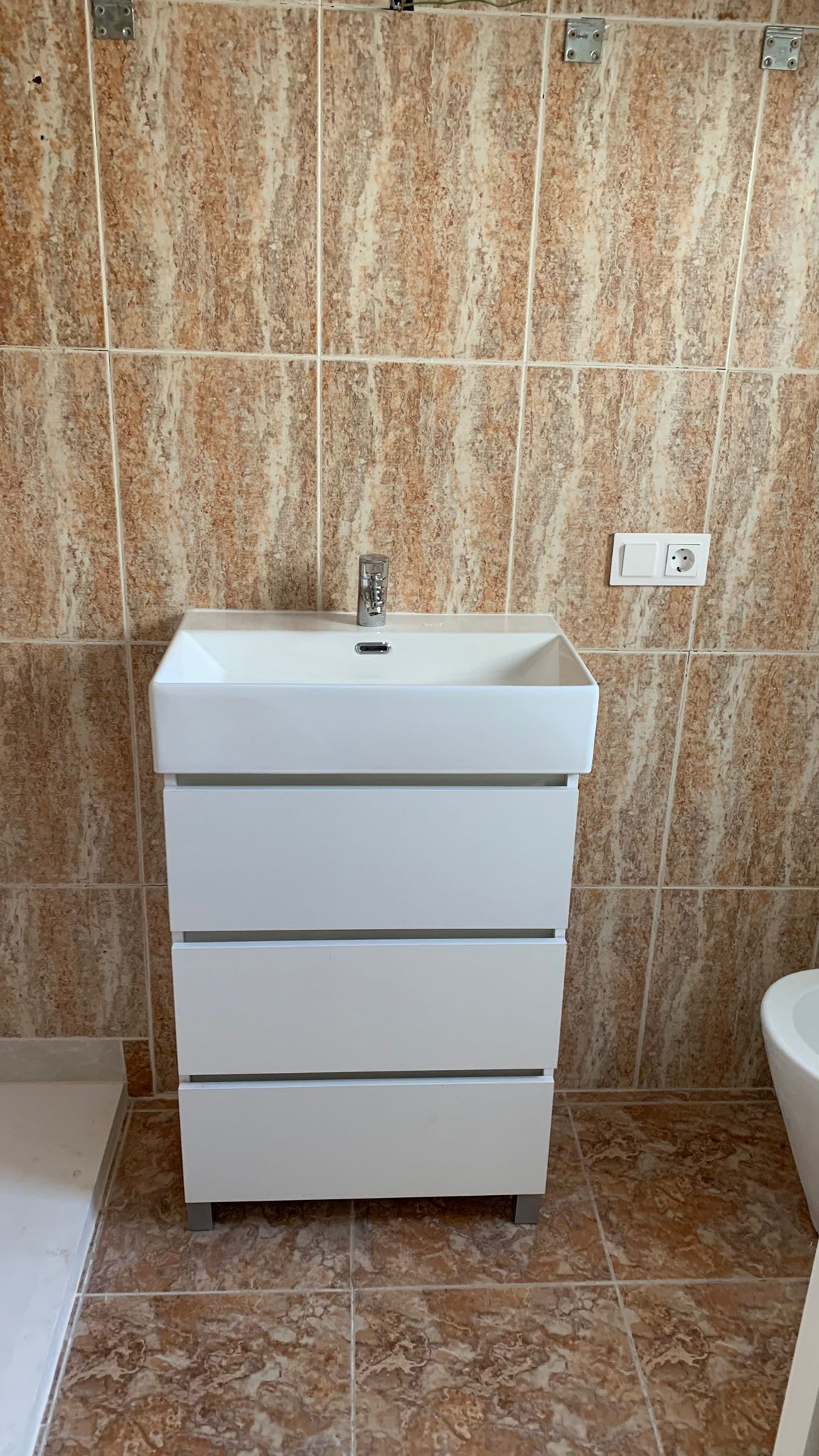 Modern white bathroom vanity featuring three drawers and an integrated sink, set against earth-toned ceramic wall and floor tiles.
