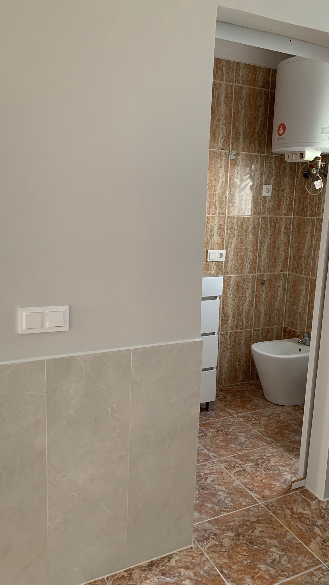 View of the bathroom featuring a bidet, electric water heater, and earth-toned tiling, seen from a hallway with grey marble wainscoting.