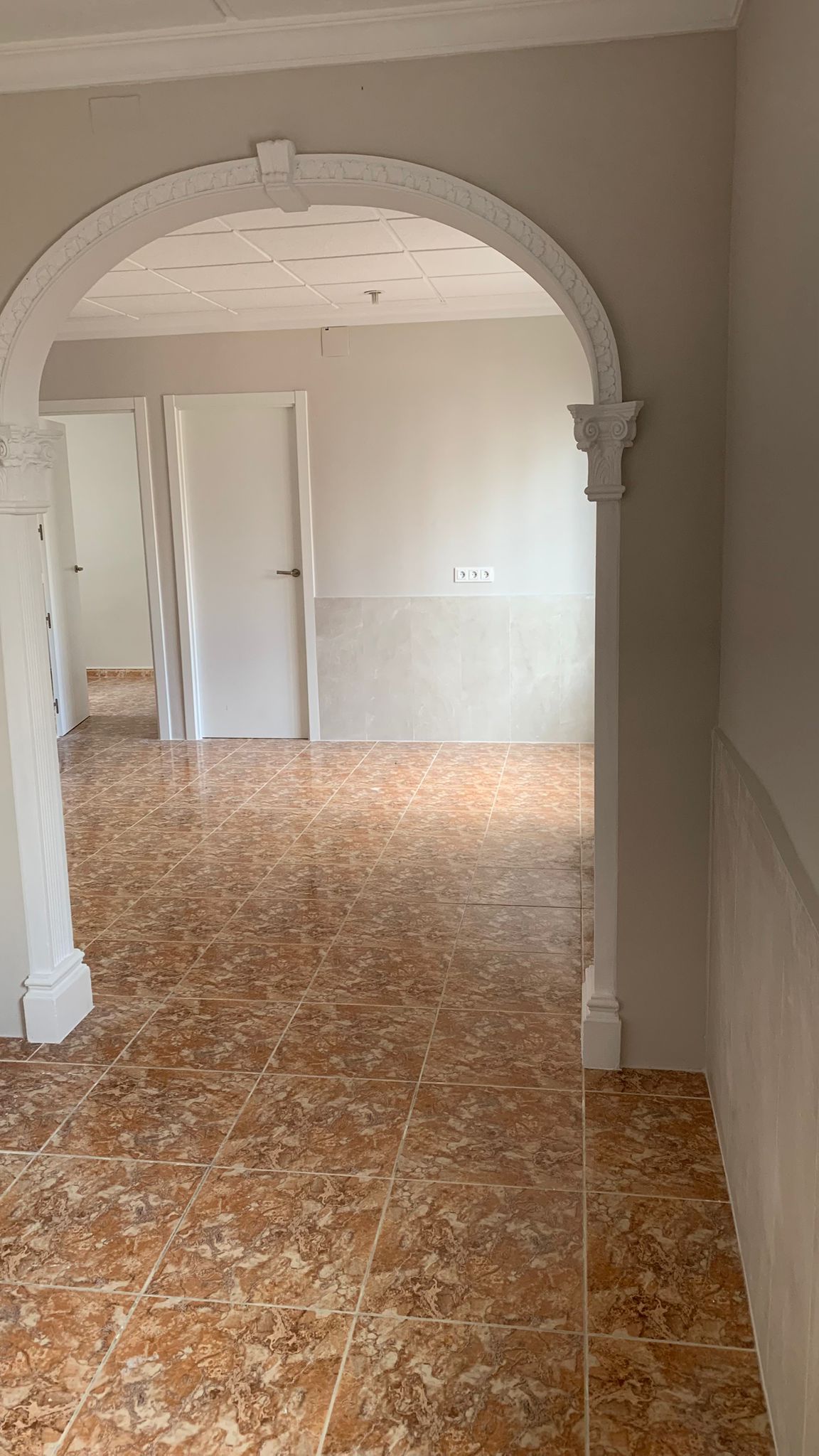 View through a white decorative archway showing brown marbled ceramic tile flooring, neutral walls, and white interior doors in a bright transition space.