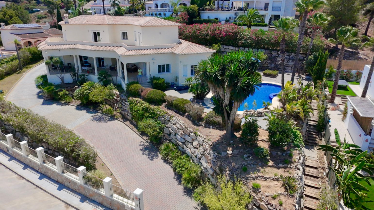 Aerial perspective of the villa featuring Mediterranean design, a private pool with palm trees, and tiered stone walls on an elevated plot in Piver, Jávea.