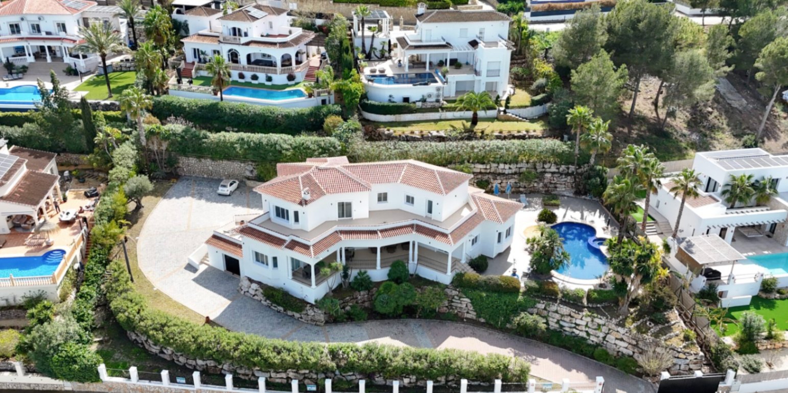 Aerial view of the main villa featuring terracotta roofs, a spacious paved driveway, a private curved pool with palm trees, and multi-level terraces set against stone walls.