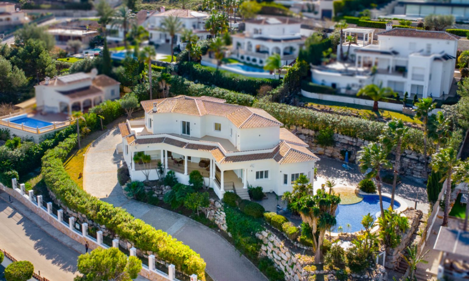 Aerial view of the villa featuring a white facade, terracotta roof, and an organic-shaped swimming pool set within a landscaped garden with stone retaining walls.