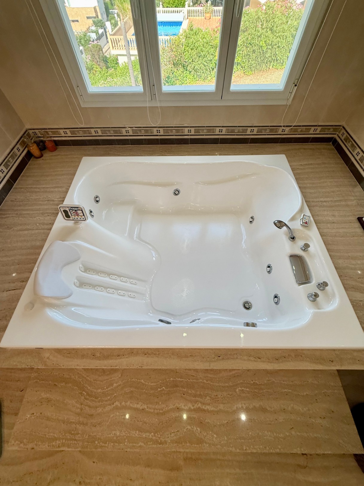Built-in hydro-massage bathtub set in natural travertine stone, featuring digital controls and a headrest, positioned under a window overlooking the garden and pool.