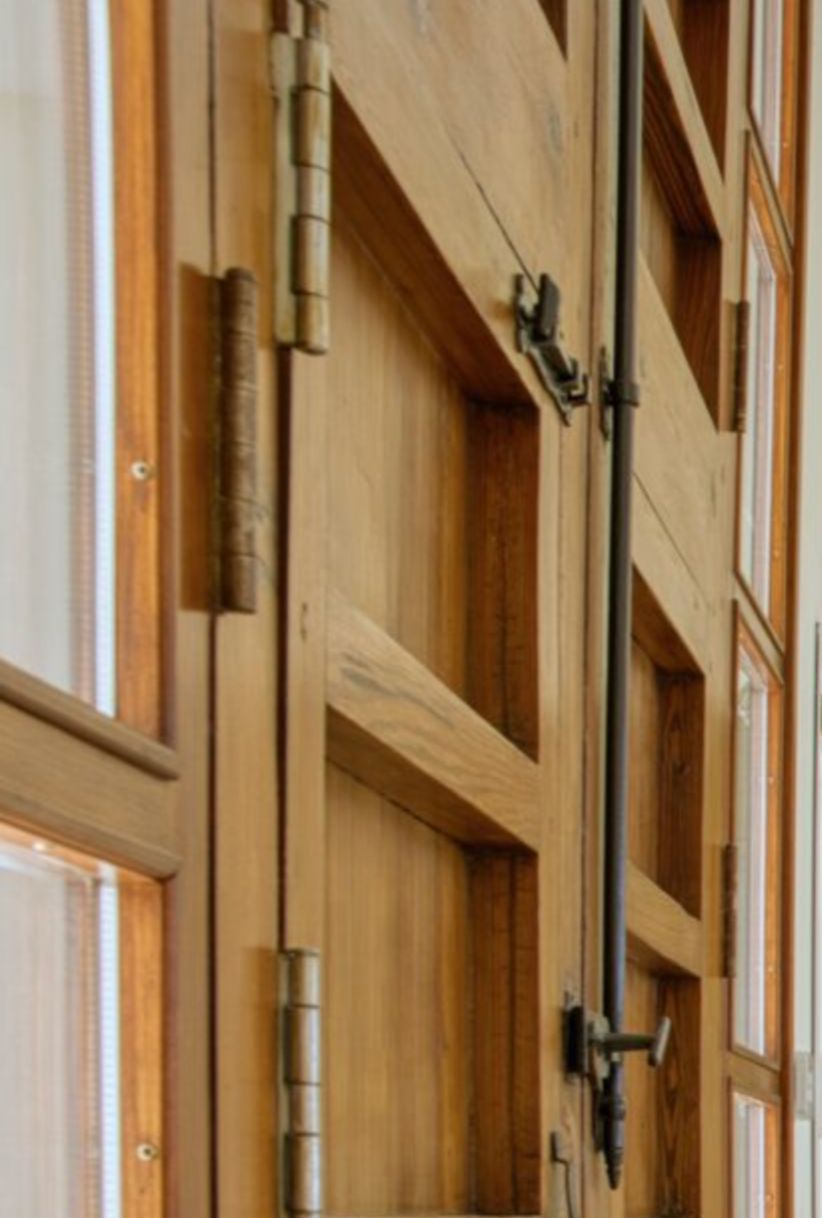 Close-up of solid wood joinery featuring traditional metal hinges and a vertical espagnolette bolt mechanism integrated with clear glass panels.