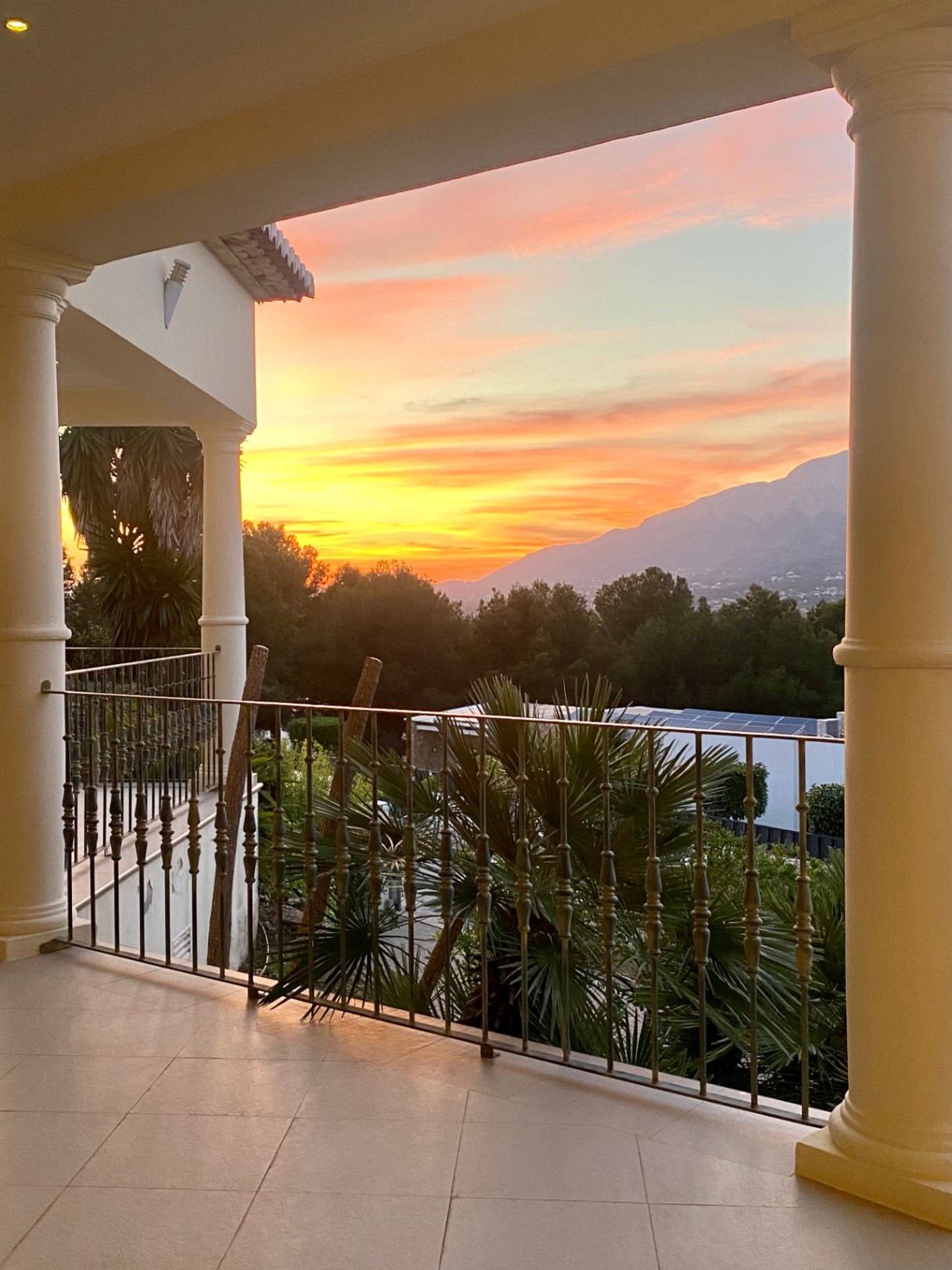 Covered terrace featuring classical columns and iron railings, showcasing panoramic sunset views over the Montgó mountain and lush Mediterranean greenery.