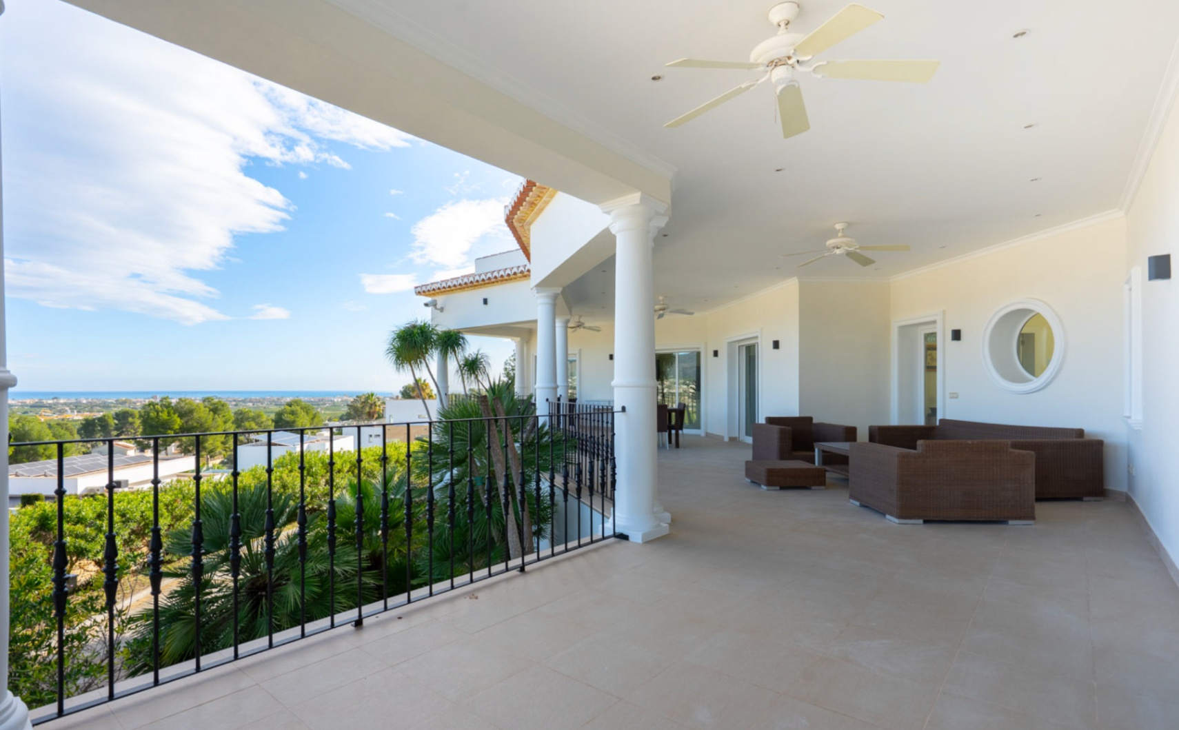 Covered terrace featuring white columns, wrought iron railings, and ceiling fans, offering a lounge area with panoramic views of the Mediterranean coastline.