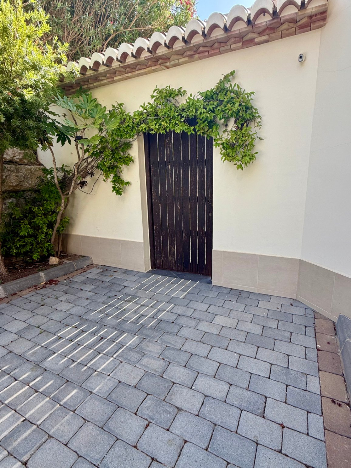Dark wood slatted exterior door framed by lush climbing greenery, featuring traditional terracotta roof tiles and a gray cobblestone paved courtyard.