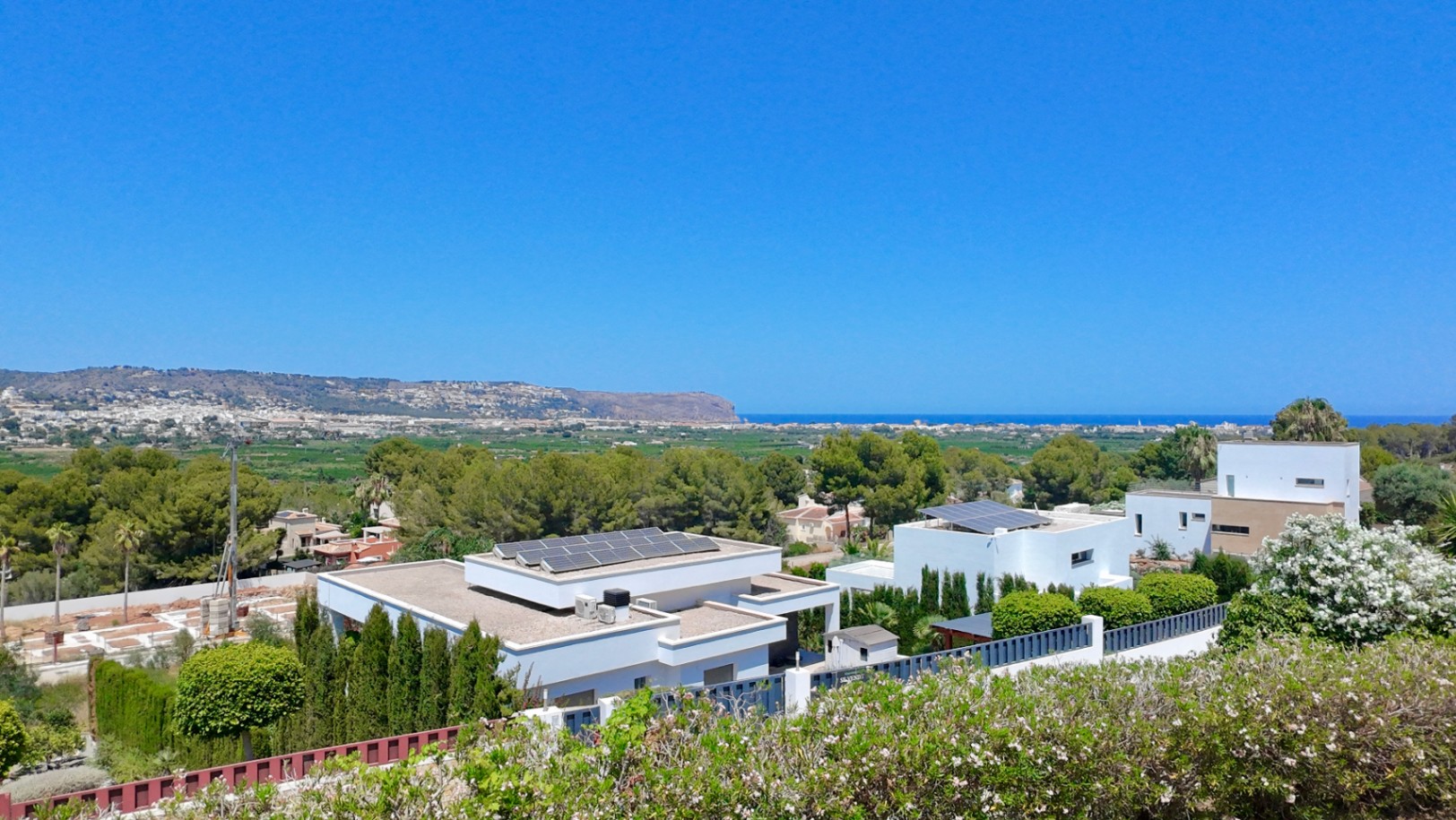 Elevated panoramic view overlooking the Mediterranean Sea and Cabo de San Antonio, featuring modern villas with solar panels amidst lush Mediterranean greenery.