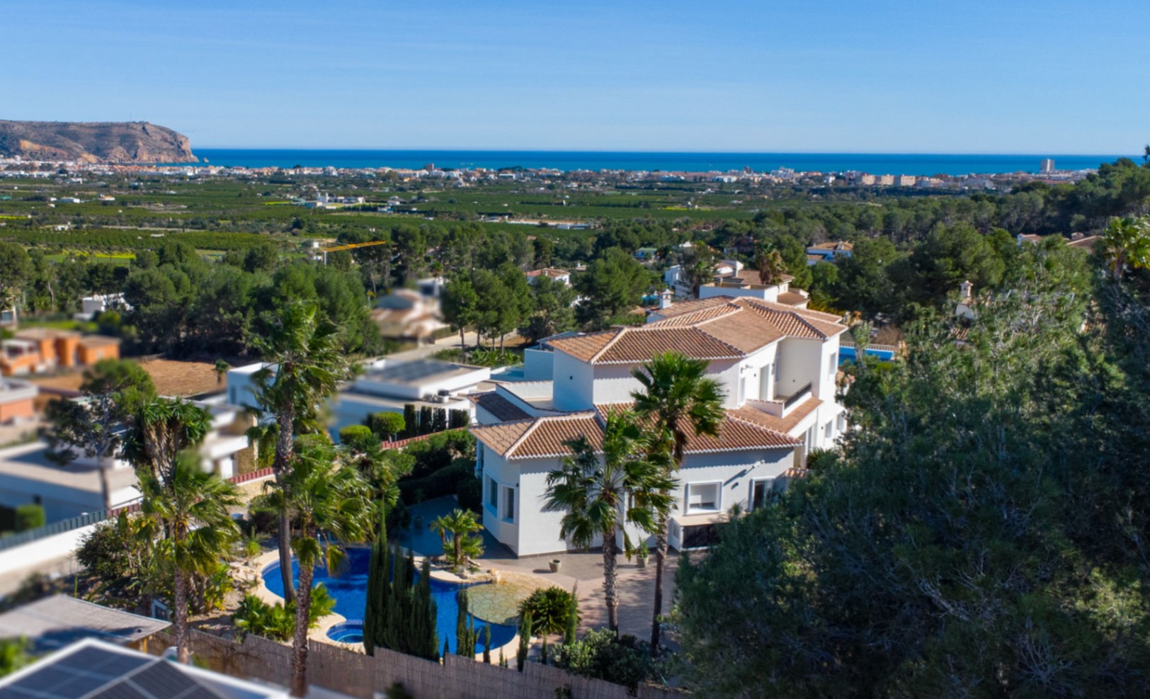 Elevated view of the white villa featuring terracotta roofs, an organic-shaped pool, and lush palm gardens overlooking the Mediterranean coastline.