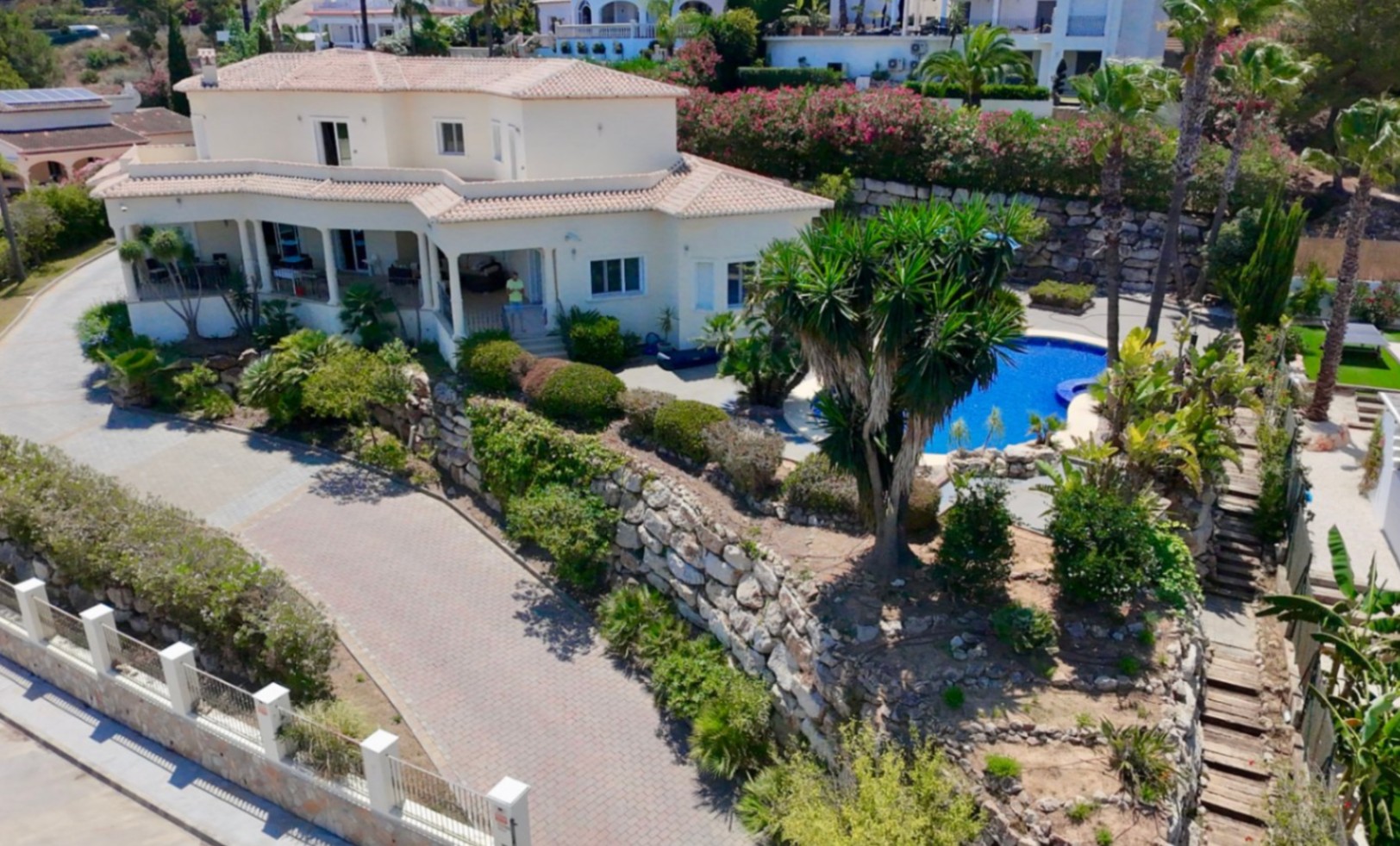 Exterior view of the villa featuring a white facade, tiled roofs, a spacious paved driveway, natural stone retaining walls, and landscaped gardens with a pool.