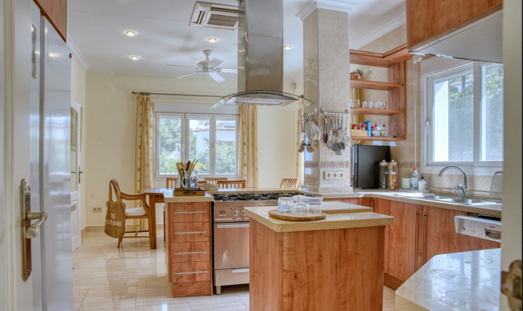 Fully equipped kitchen featuring a central island with a stainless steel range, stone countertops, and wooden cabinetry. Includes recessed lighting and an integrated dining space.
