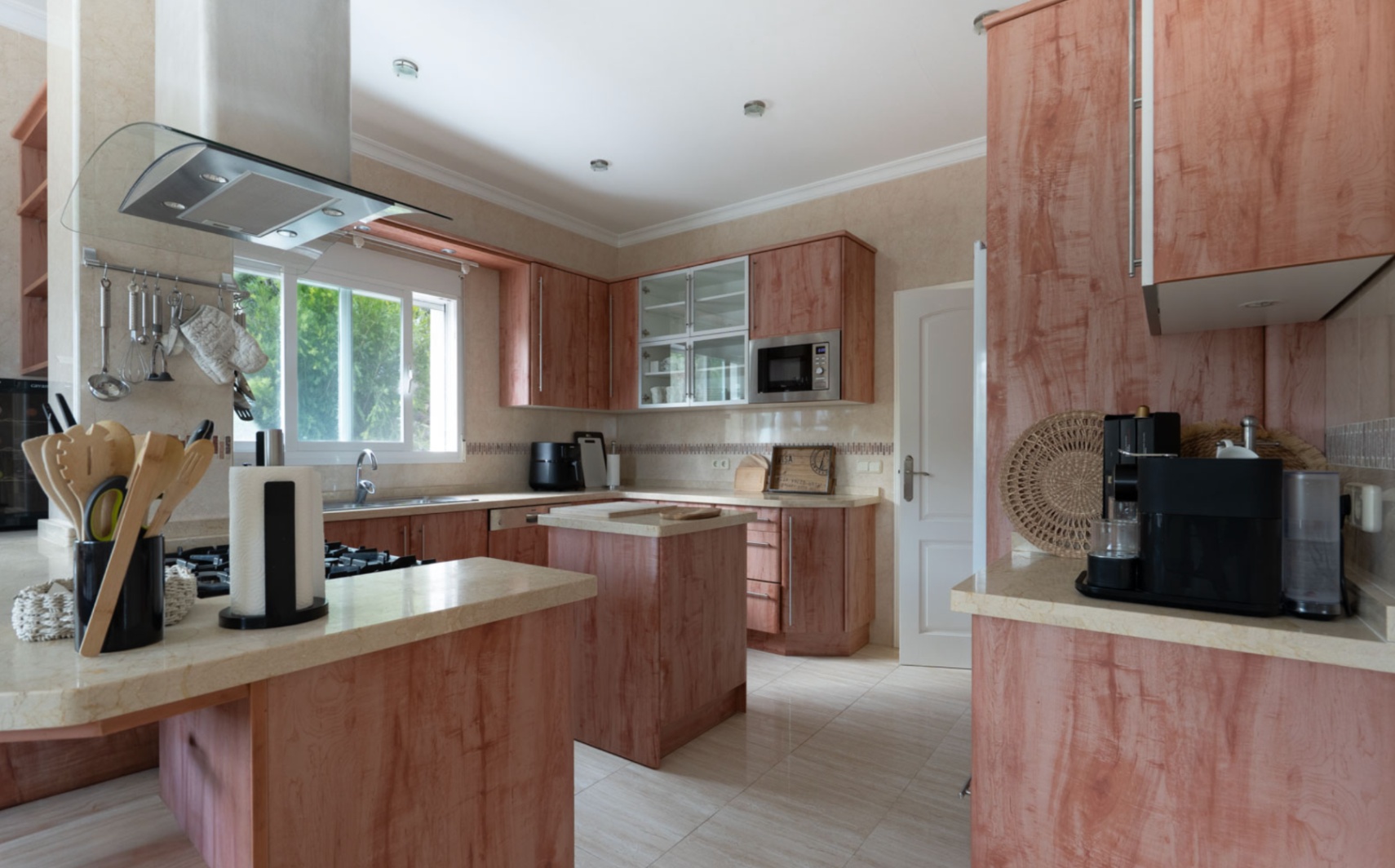 Fully equipped kitchen featuring warm wood cabinetry, beige stone countertops, and a central island. Includes integrated stainless steel appliances and a large window facing the garden.