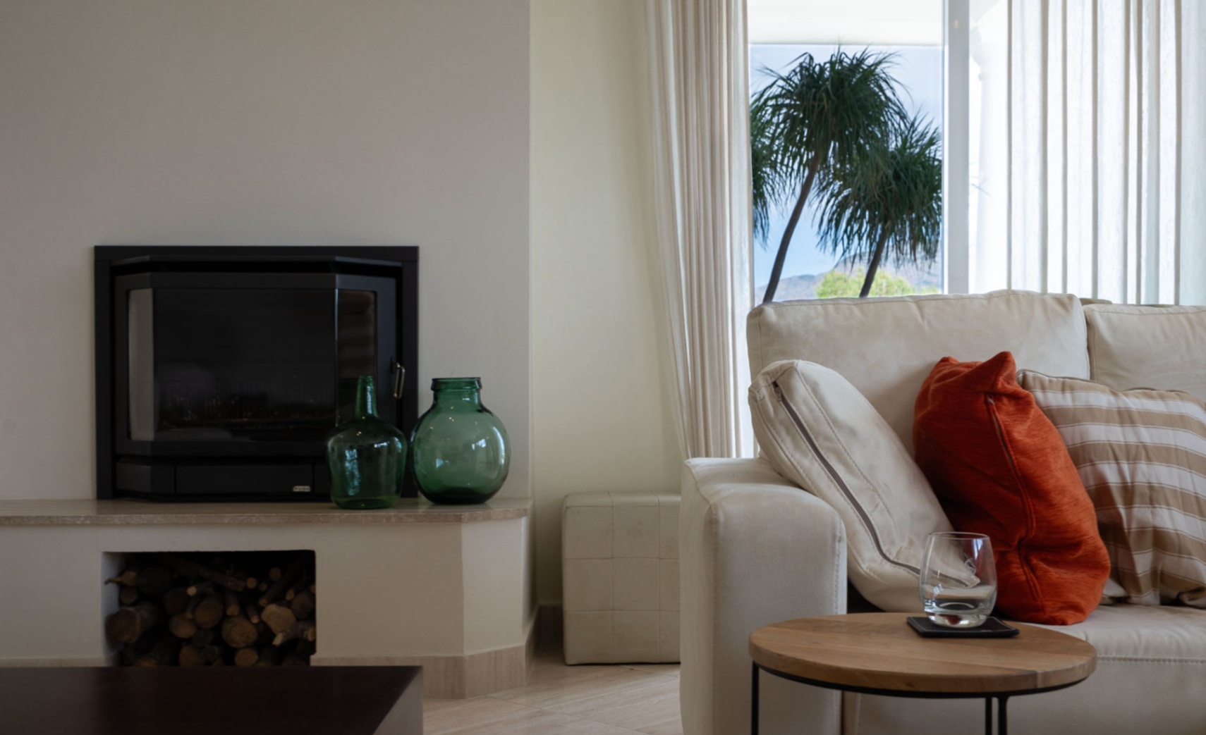Living room featuring a black built-in fireplace with wood storage and views of palm trees. The space is finished with neutral tones and natural light.
