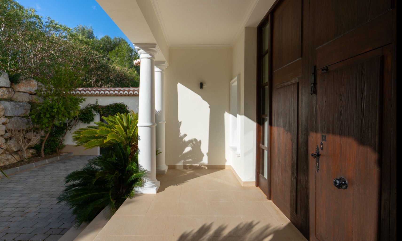 Main entrance featuring a solid dark wood door and classical white columns. The covered porch overlooks a stone-paved driveway and lush Mediterranean greenery.