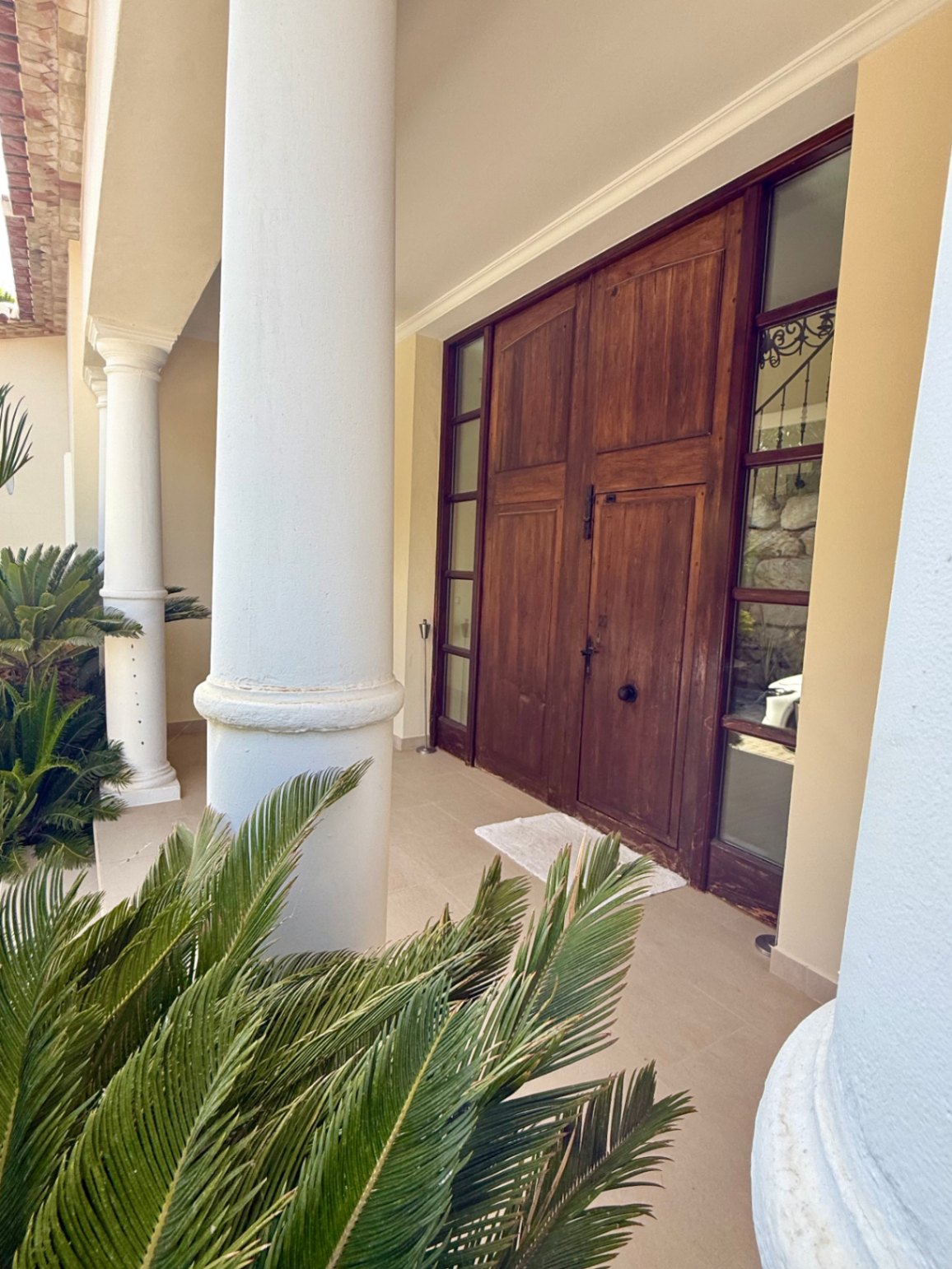 Main entrance featuring a solid dark wood double door with side lights, framed by white Tuscan columns and lush Cycas plants on a shaded porch.