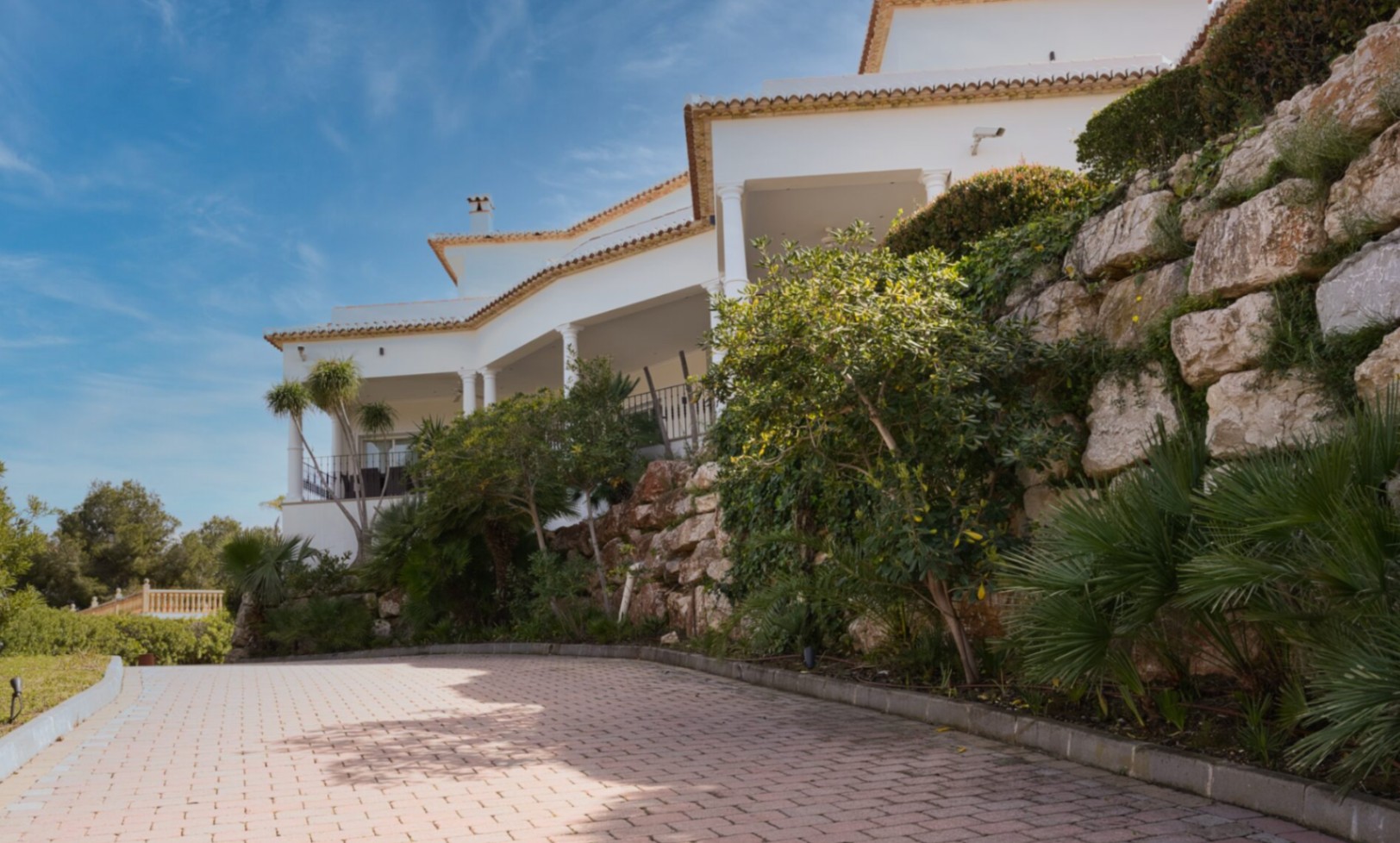 Mediterranean villa exterior featuring white columns, covered terraces, and a paved driveway bordered by a natural stone retaining wall and lush greenery.