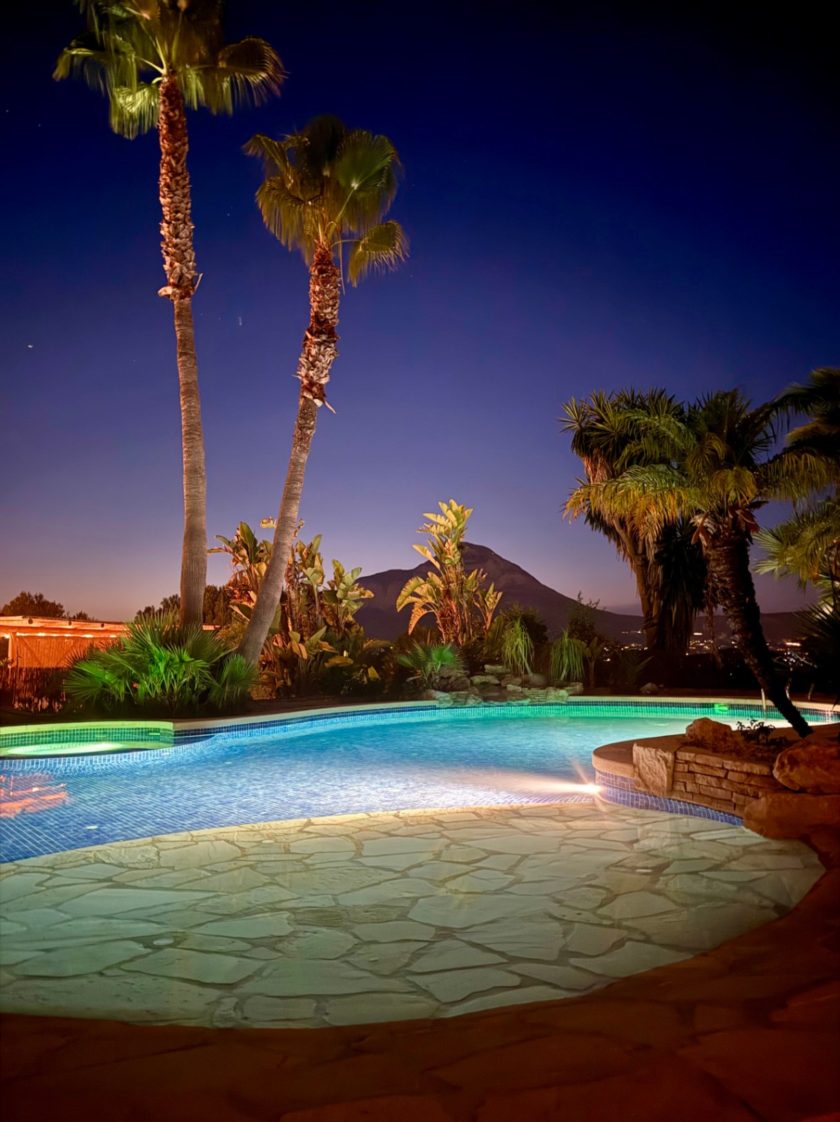 Night view of the illuminated swimming pool featuring natural stone paving and a backdrop of the Montgó mountain framed by palm trees.