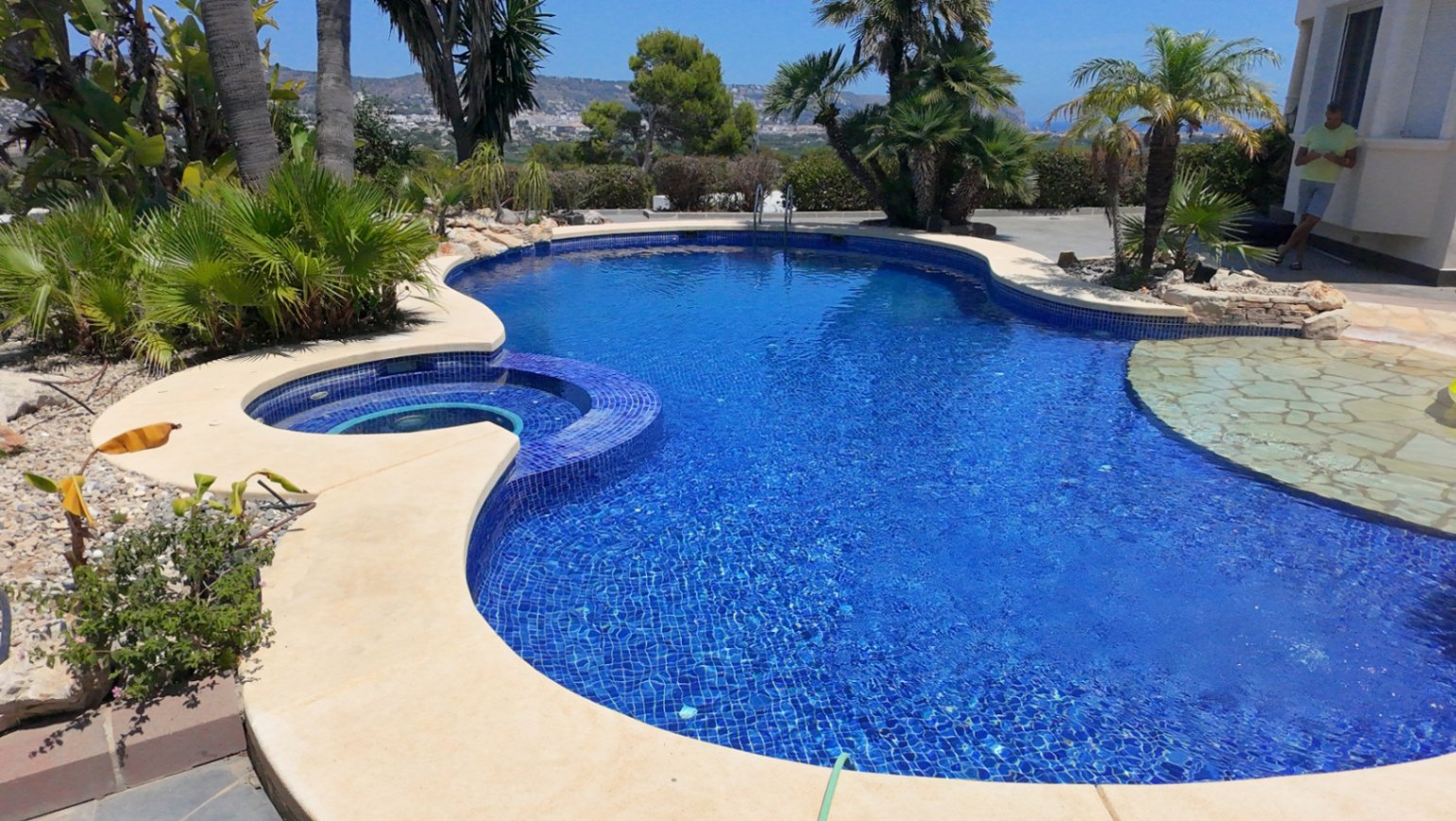 Organic-shaped swimming pool featuring blue mosaic tiling, an integrated circular spa, and a natural stone entry area, set amidst mature palm trees.