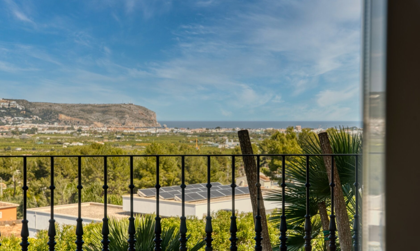 Panoramic view from the upper floor overlooking the Mediterranean Sea and coastal cliffs, framed by a black wrought iron railing and native palm trees.