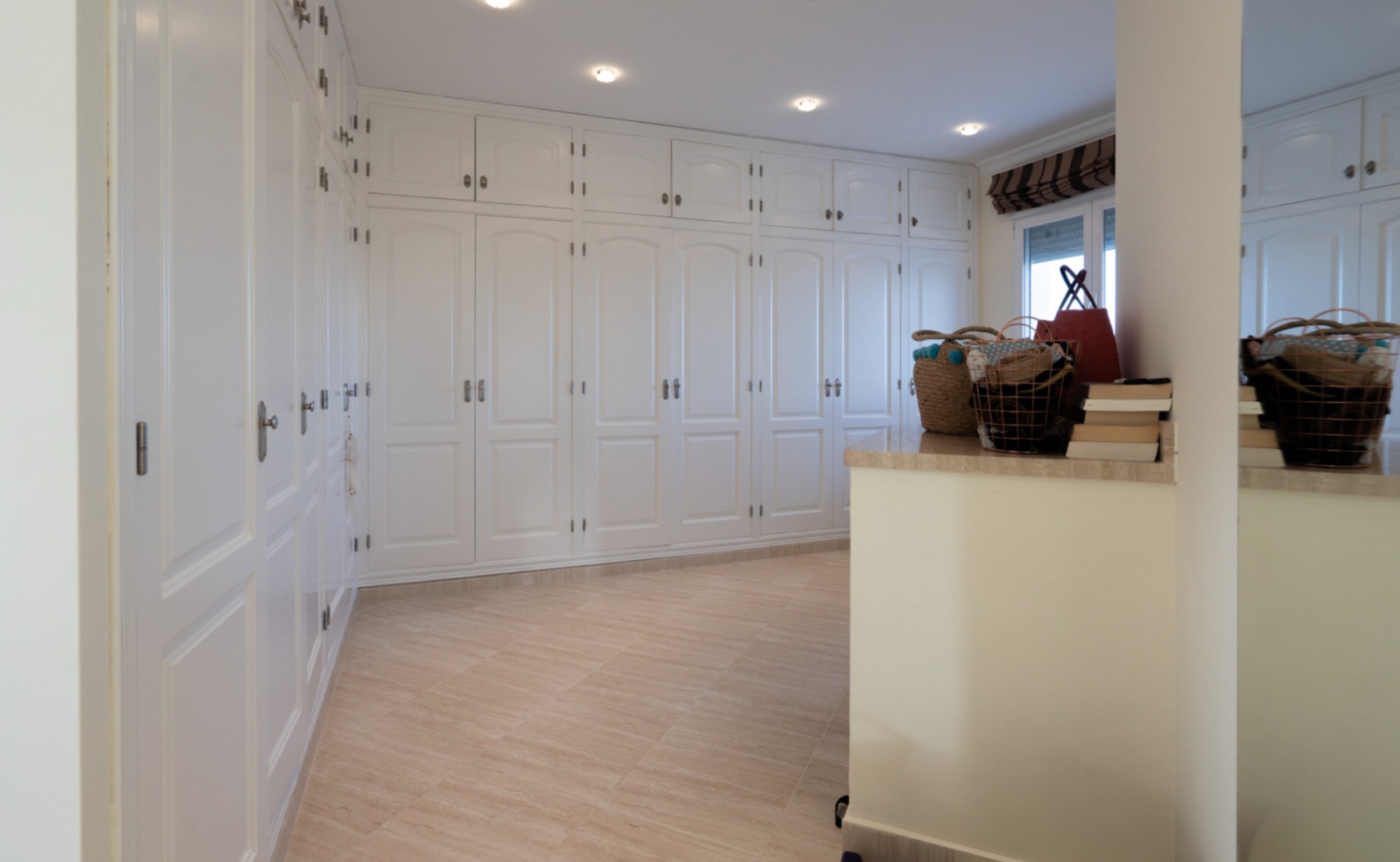 Spacious walk-in closet featuring floor-to-ceiling white fitted wardrobes with arched panels and recessed lighting. Polished tile flooring enhances the bright space.